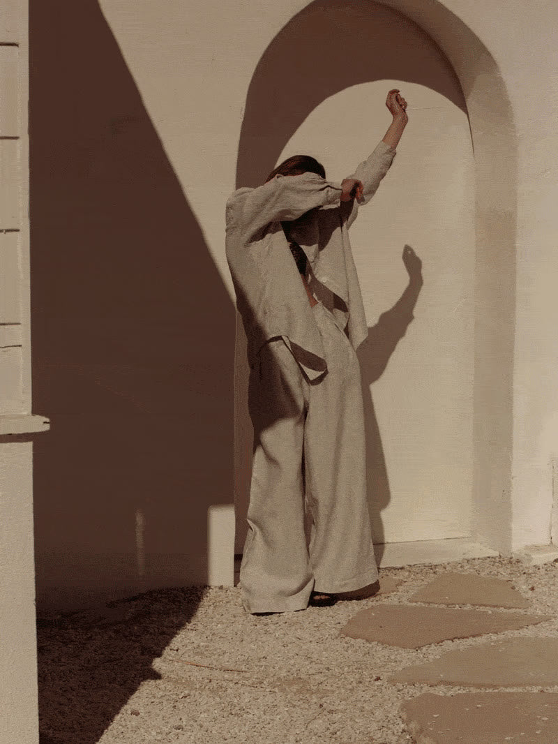 A woman in a relaxed linen outfit moving in warm sunlight beneath an archway.